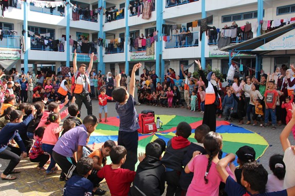 En folkmassa med barn står runt en Röda Korset volontär utomhus. Barnen hoppar och dansar i en aktivitet som hålls.
